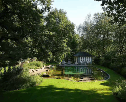 Blick auf einen organisch angelegtem Schwimmteich mit Gartenhaus, Rasen und altem Baumbestand