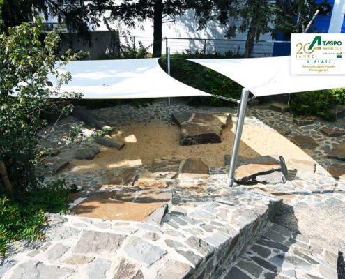 Das Kita-Außengelände mit Naturstein, Totholz und kleinen Felsenhängen Sonnensegel und Fläche aus Naturstein
