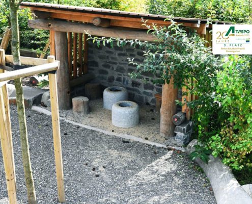 Naturnaher Spielbereich mit Sitzplätzen aus Holz und Naturstein Überdachtes Spielelement aus Holz und Grauwacke