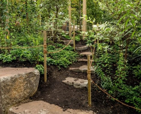 Ein schmaler Weg mit Natursteinstufen, eingefasst von Holzpfosten und Seilen.