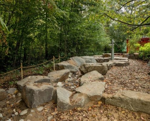 Eine natürliche Spiellandschaft mit großen Felsen, Laub und einem Wasseranschluss, umgeben von dichter Vegetation.