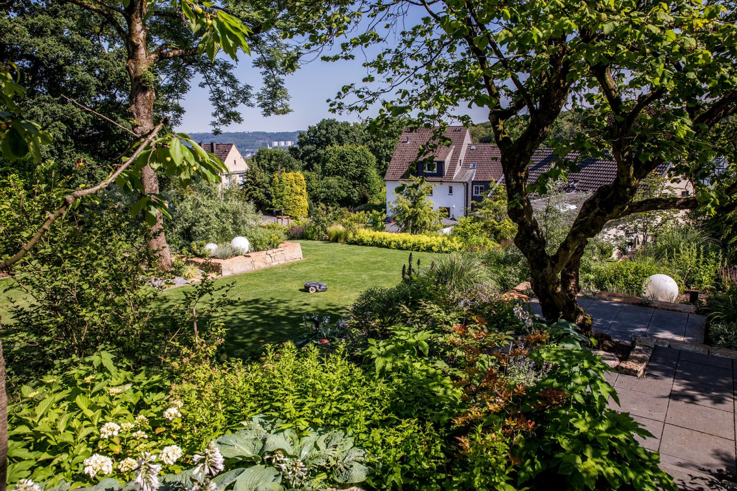 Blick auf weitläufigen Hanggarten mit großer Rasenfläche, üppig bewachsenen Beeten und Terrasse