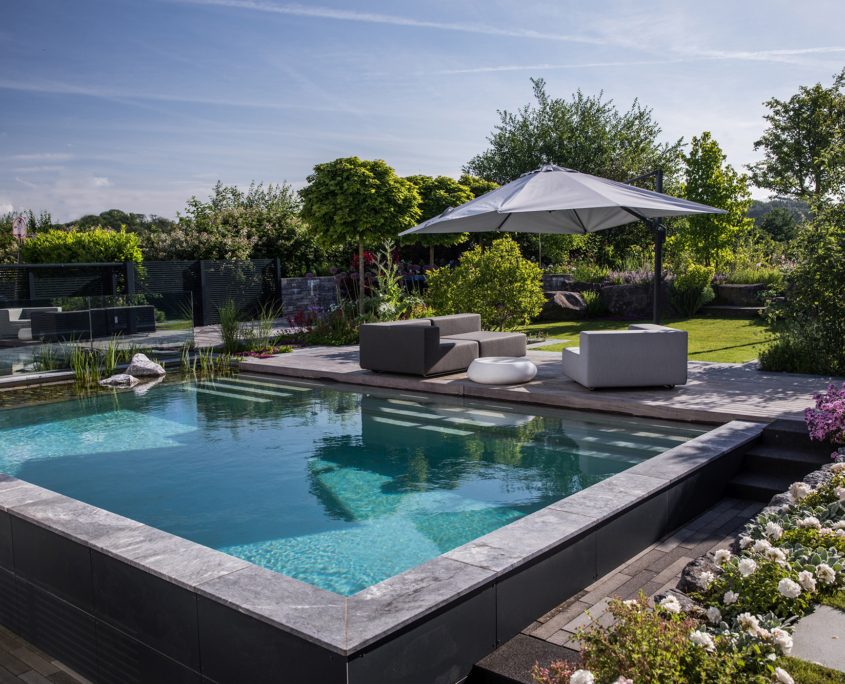 Naturpool neben einem Deck mit Loungemöbeln inmitten üppiger Bepflanzung
