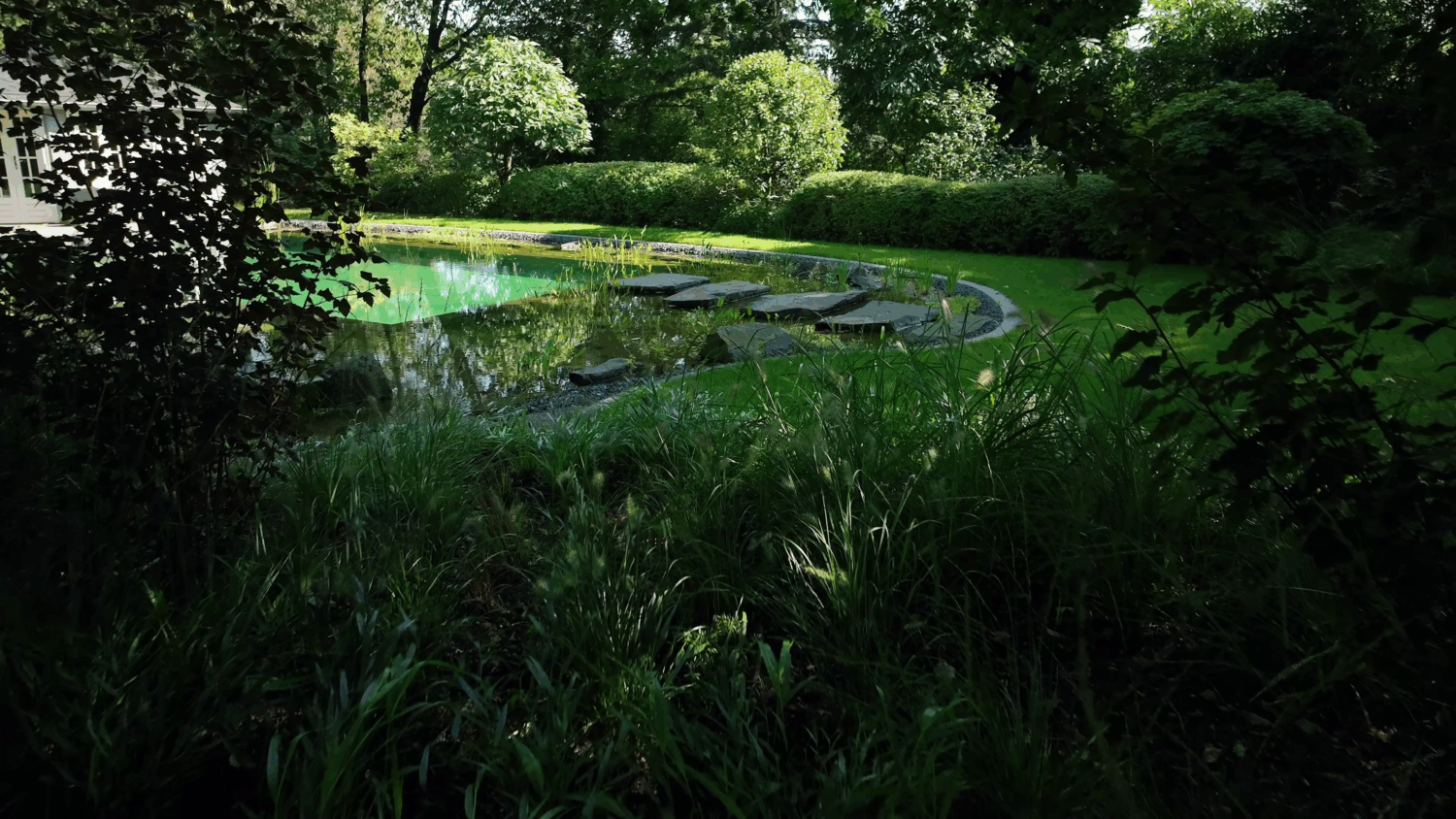 Tiefgrüner Garten mit üppiger Bepflanzung und großen Natursteinen rund um einen Schwimmteich